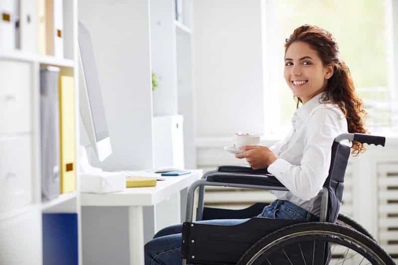 young woman in wheelchair at computer holding cuyp of coffee.
